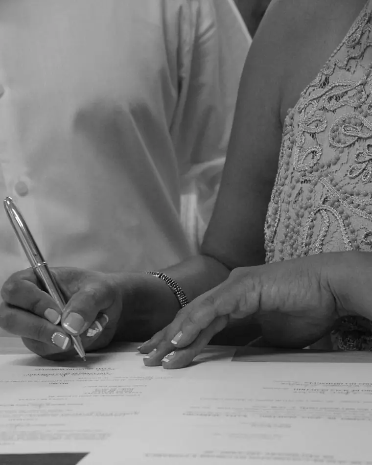 Black and white photo of a couple's hands at a table, one signing a document with a pen