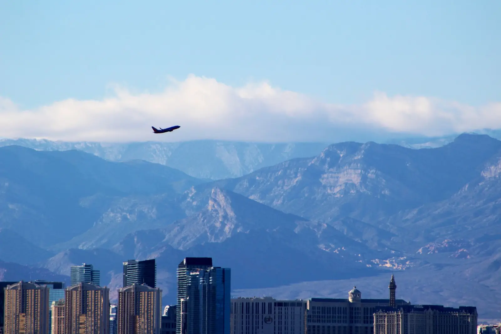 A plane climbing out over the Las Vegas skyline, the mountains and a bank of white cloud behind it
